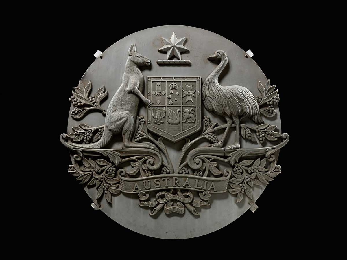A metallic Australian coat of arms mounted on a large metal disc with four brackets attached. The coat of arms is cracked across the feet of the emu, between the emu and the shield, and through the wattle branch behind the kangaroo.