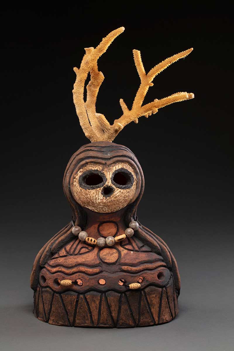 A red raku clay sculpture roughly in the shape of the torso and head of a human. It has coral for antlers and is painted mainly in shades of brown. The face is painted white with black borders around the eyes and nose. There are two leather cords protruding from the torso threaded on which are two wooden beads. Hanging from a leather cord around the neck are six round and two long wooden beads. - click to view larger image