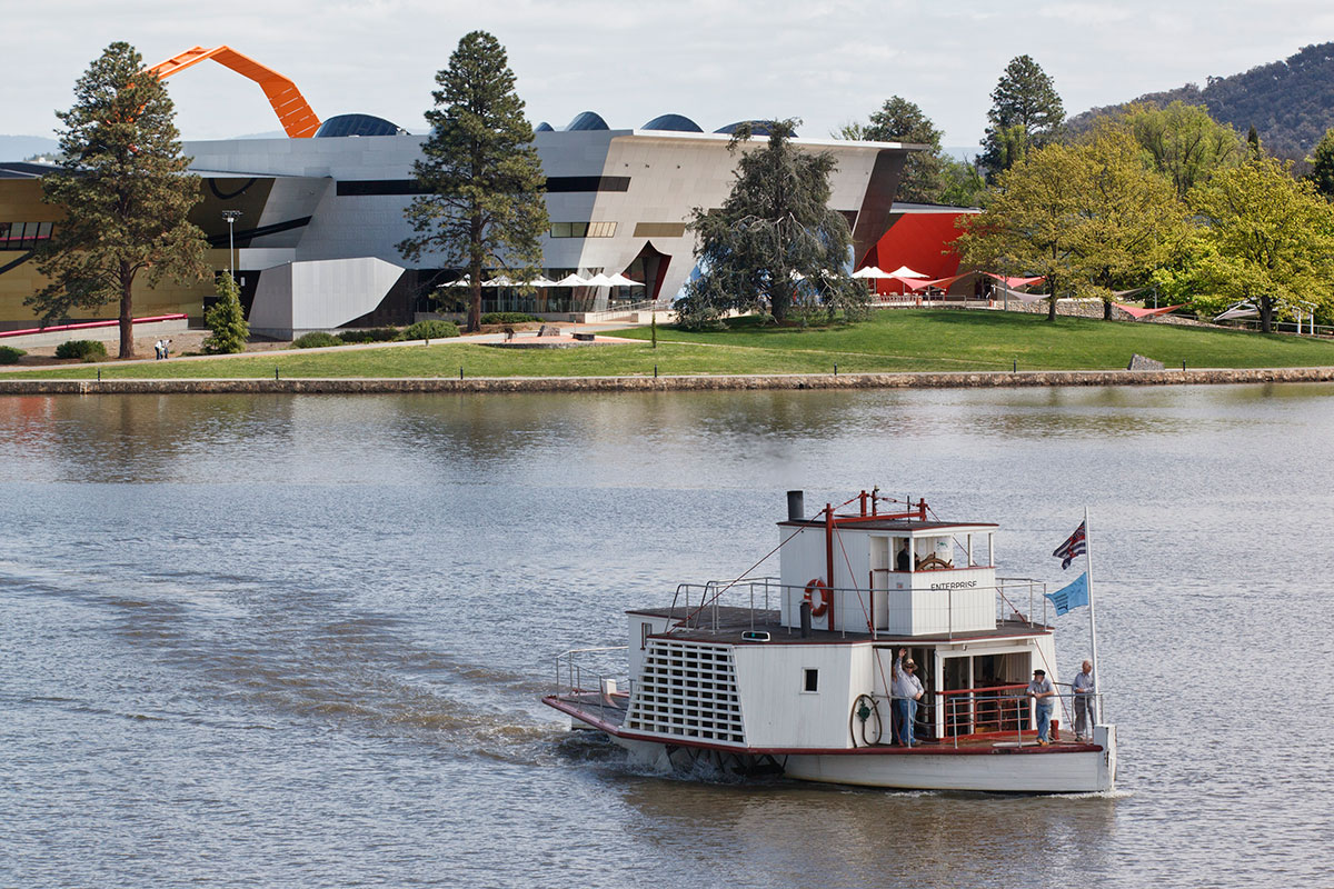 A view of a paddle steamer on a lake, with a modern museum building on the shore.