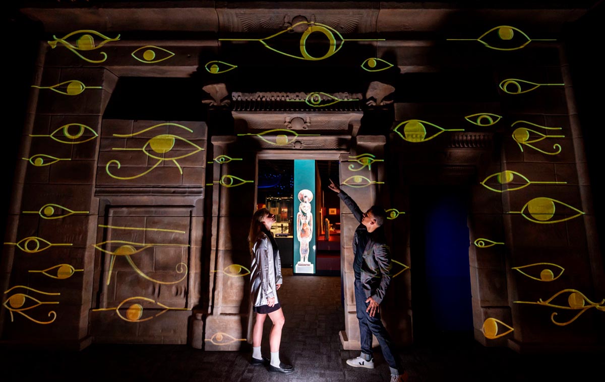 Two visitors inside a darkened exhibition space, looking at hieroglyphs projected on a replica temple wall. 