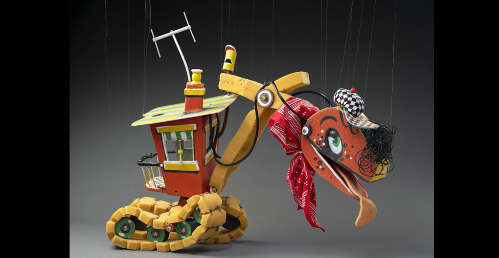 A puppet named Bill Steamshovel, in the form of an excavator. Bill Steamshovel consists of a red, yellow and green wooden control room connected to which are foam tread tyres and a foam neck. At the end of the neck is a red and yellow excavator head with a checkered hat and hair made from strands of black rubber. Tied around the neck is a red and white handkerchief.