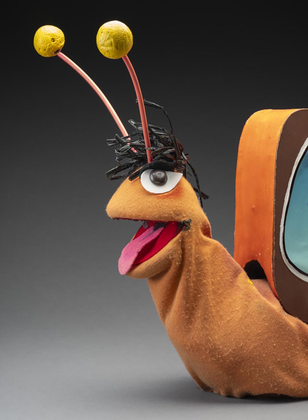A puppet of a snail named Gus. Gus has an orange body, a tuft of black hair, orange and yellow antennas, brown eyes and a pink felt tongue. Attached to its back is a television box with two knobs, a screen featuring an image of Gus’s head surrounded by a gold frame, and printed text that reads ‘GUS/TV’. Attached to the top of the television is a yellow flowerpot with a red flower, sitting atop a doily.