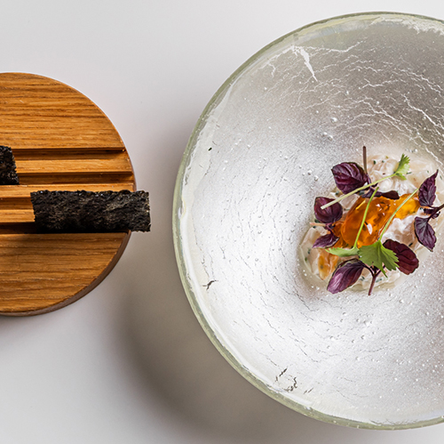 Photograph of a beautifully set tableware and food. A portion of food sits neatly in a bowl, to the left of the bowl with is smaller wooden circular bowl. Above the food sits a wine glass and water glass.