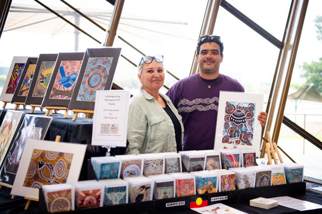 Two people stand behind a table displaying brightly coloured First Nations prints and cards. - click to view larger image