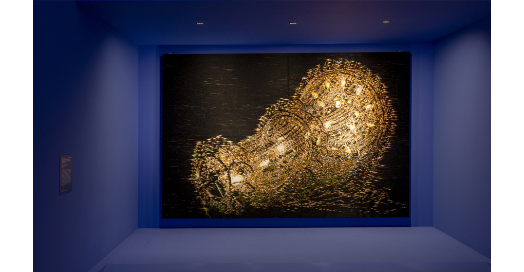 A large piece piece of needlework depicting an elaborate three tiered chandelier, illuminated but laying on its side, not suspended. The background is a field pitch black on which the image of a chandelier is constructed with long stitches of fine, golden coloured thread.