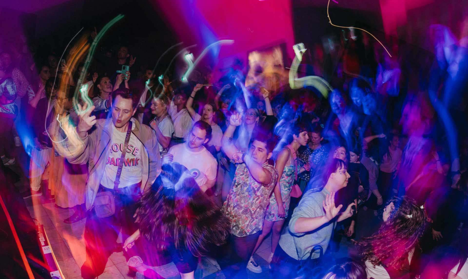 A photograph of a museum party with blue and pink lights. The crowd is dancing and energetic.
