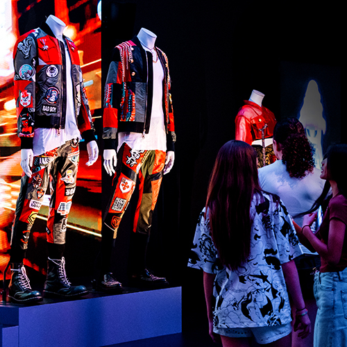 Photograph of three people looking at a Korean fashion display in the gallery. The display is of mannequins wearing colourful Korean inspired street clothes. Bind the mannequins are bright screens with videos playing.