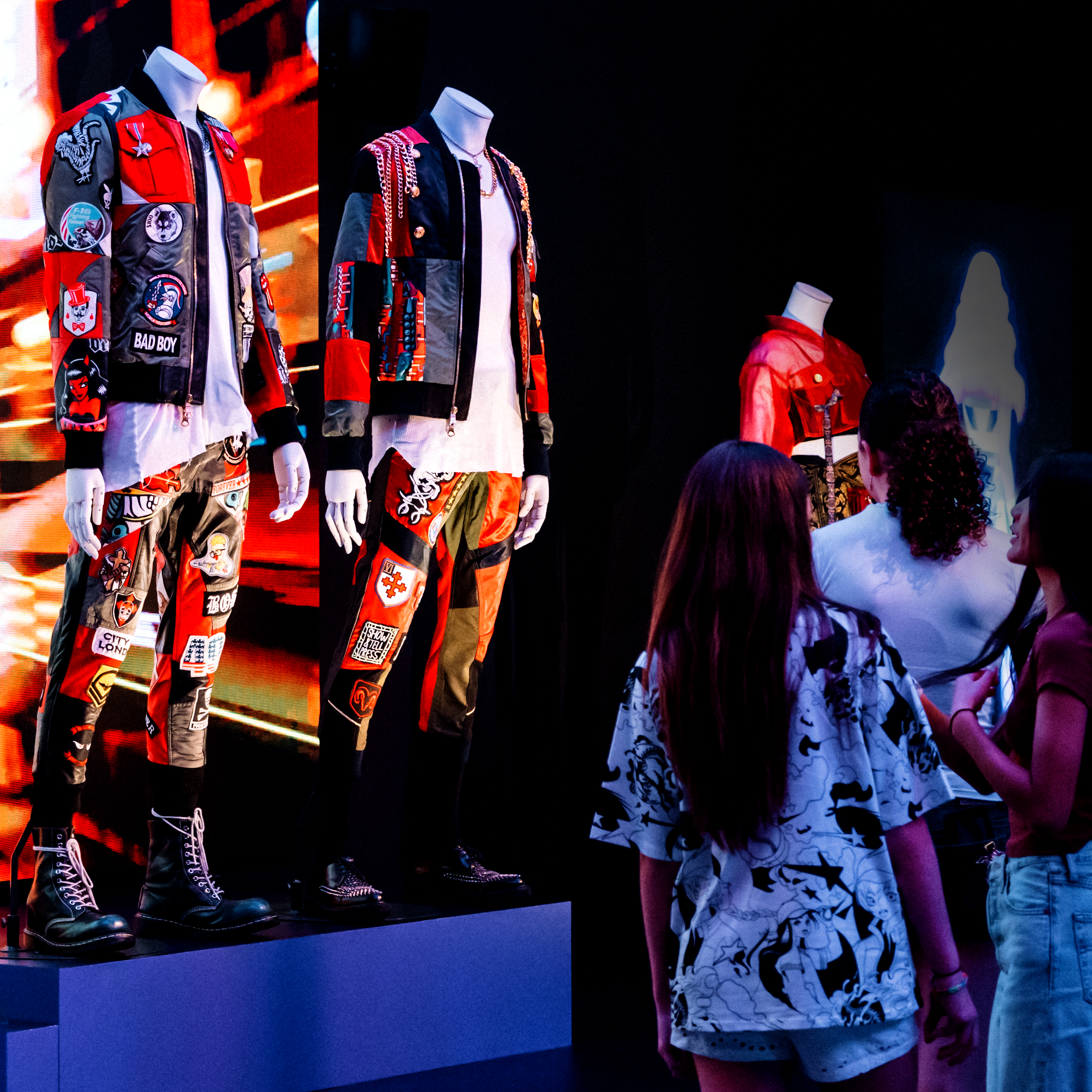 Photograph of three people looking at a Korean fashion display in the gallery. The display is of mannequins wearing colourful Korean inspired street clothes. Bind the mannequins are bright screens with videos playing.