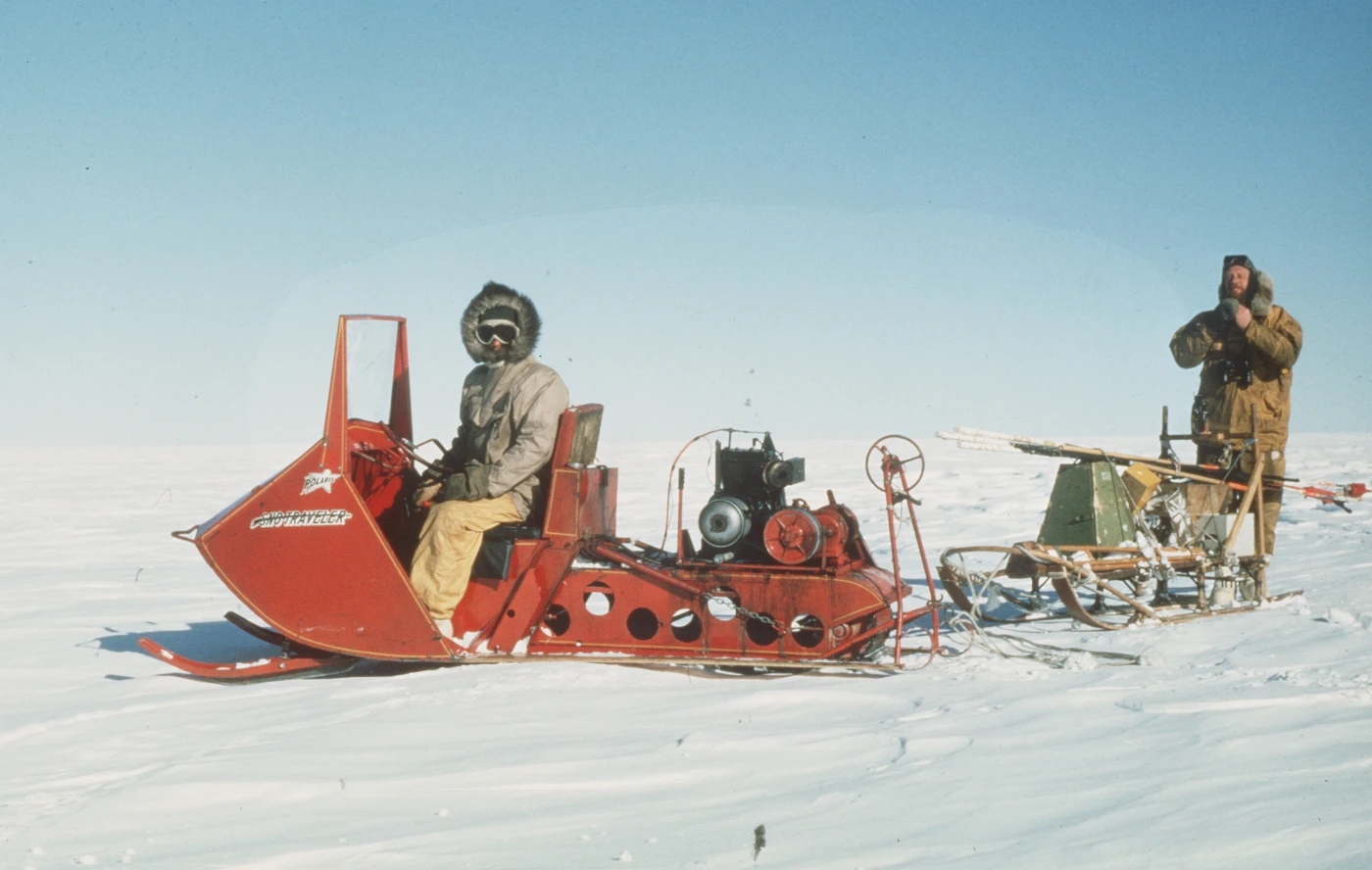 Image showing two men in an Antarctic landscape. One sits in the open cabin of a bright orange oversnow vehicle, on tracks. The second person stands at the back of the vehicle, which tows equipment on a sled.
