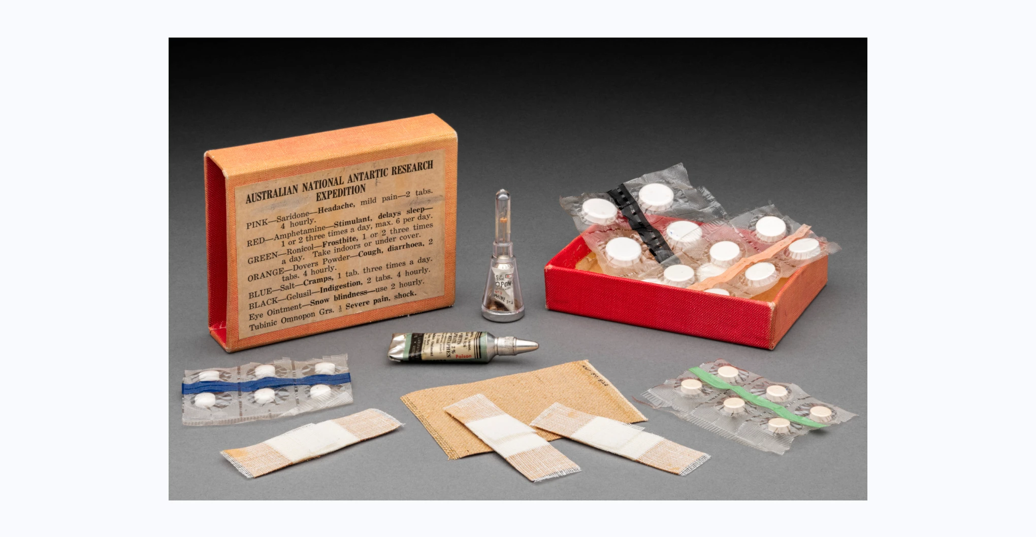 An Australian National Antarctic Research Expedition first aid kit. The kit is housed in a red and pink rectangular cardboard box with a slide-out tray, and its contents are described on a printed label on the lid. Some of the described contents are missing from the kit. Included are four plastic sachets of tablets: Ronicol for frostbite, Gelusi for indigestion, Saridone for headaches, and salt for cramps. Also included are a tube of eye ointment for snow blindness, an ampule of Tubinic Omnopon Grs for severe pain, three Elastoplast strips and one adhesive patch. Along the top of the label is faded handwritten text in black ink that reads 'SAMPLE ONE [...?] MEDICATION'. Handwritten in pencil on the bottom is 'MB Pittman / MB 4411'.