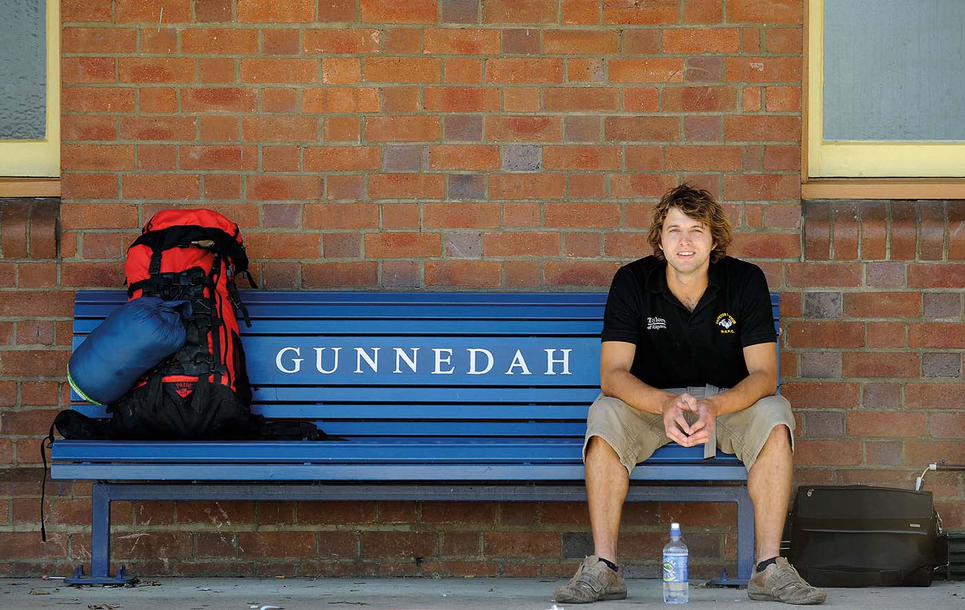 Landscape image showing a man sitting to the far right of a blue, slatted railway bench. 'GUNNEDAH' is painted in white at the centre back of the bench. The man wears a black open-collared T-shirt and khaki shorts and sits with his forearms resting on his thighs. A red and black backpack and a blue sleeping bag sit at the opposite end of the bench. Reddish-coloured bricks form a backdrop.