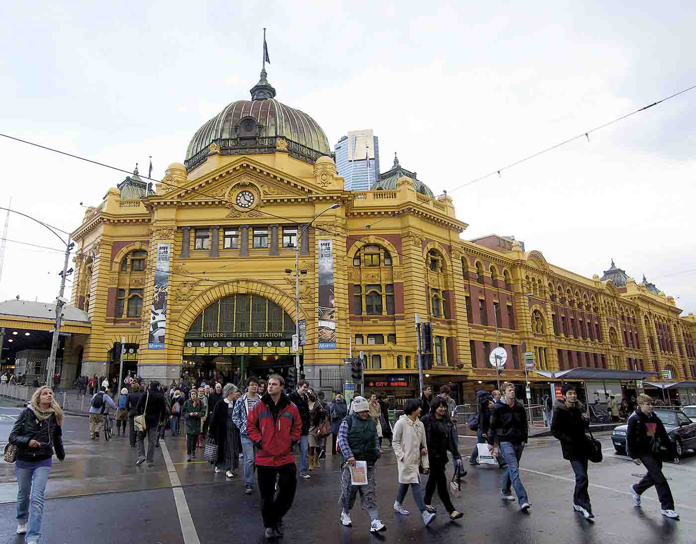 A crowd of people walks across a paved road outside Flinders Street Station, an Edwardian baroque building with a golden-coloured and red brick facade. Tram lines extend in various directions above the road.