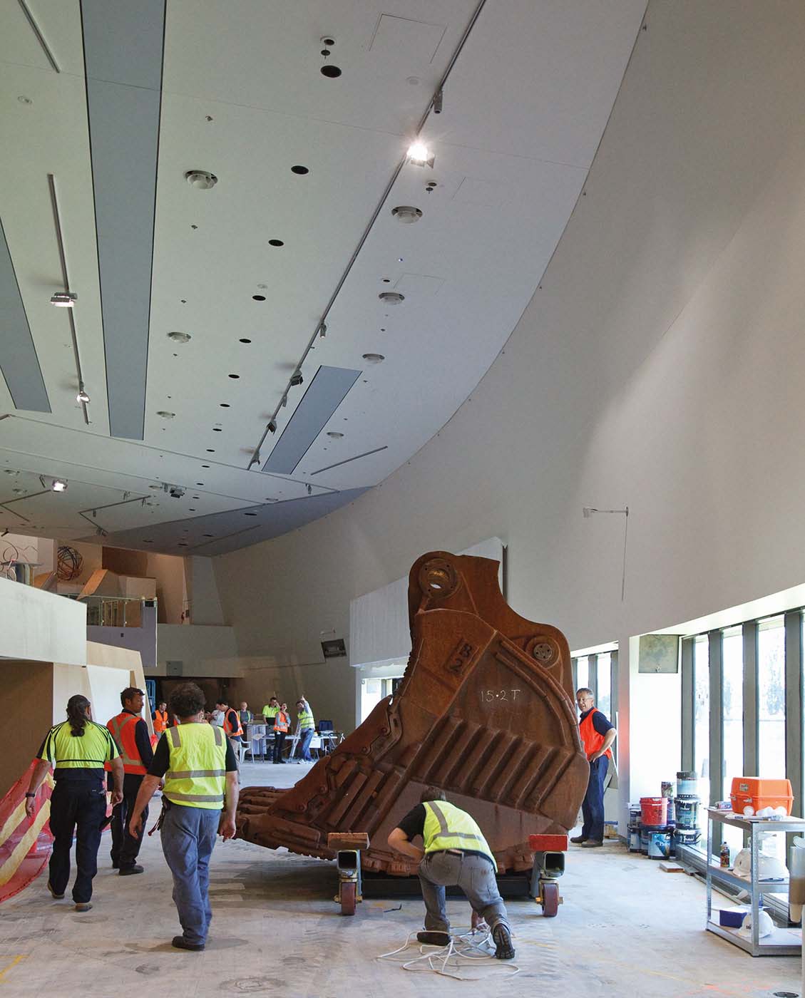 Side view of a rusty, concave-shaped iron bucket from a mining excavator. '15.2T' is spray-painted in white on the side. The bucket rests on a wheeled trolley and sits in a large, caverous gallery. Several workers in flourescent-coloured vests stand near the bucket and others gather in the distance.