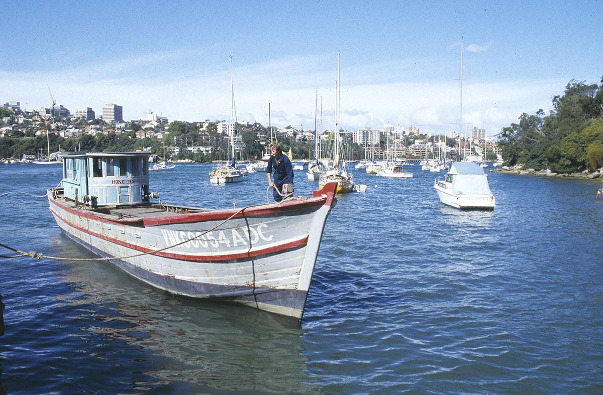 Colour photograph of a person on a fishing boat in a harbour with the name 'Hong Hai' painted on the outside.