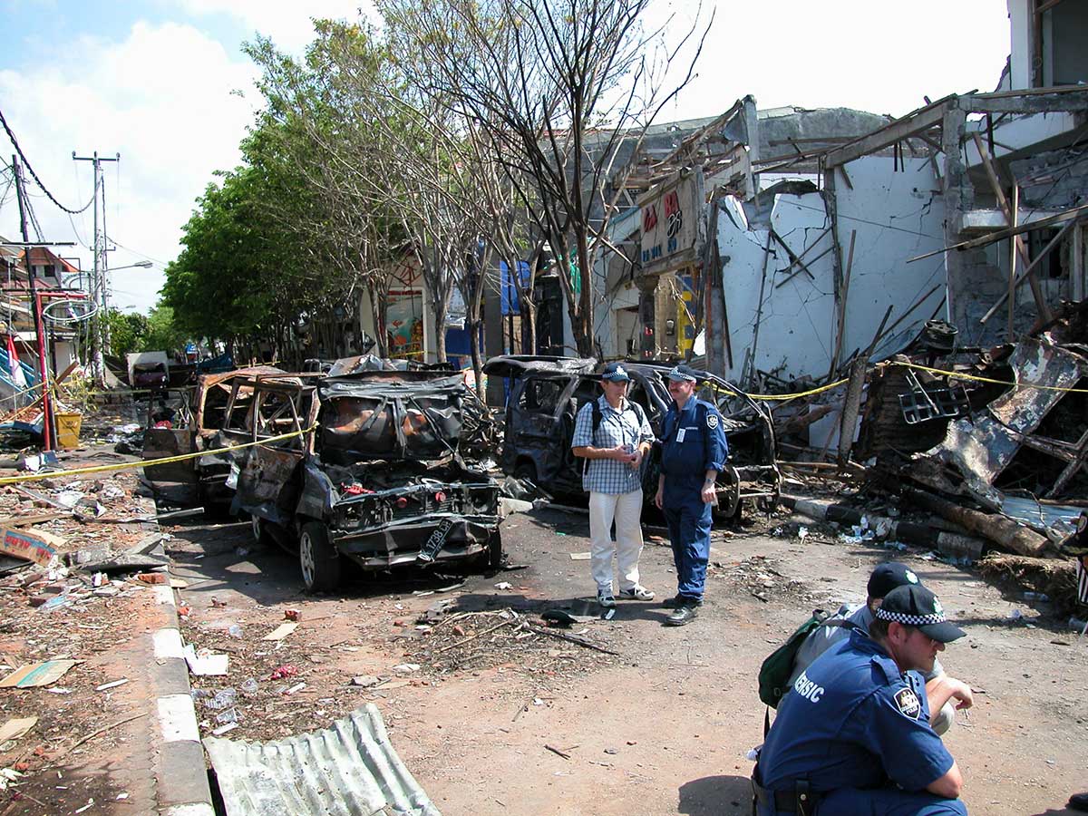 Four Australian Federal Police officers, two in uniform, confer in the street. Behind them are the burnt-out remains of vehicles and a wrecked building