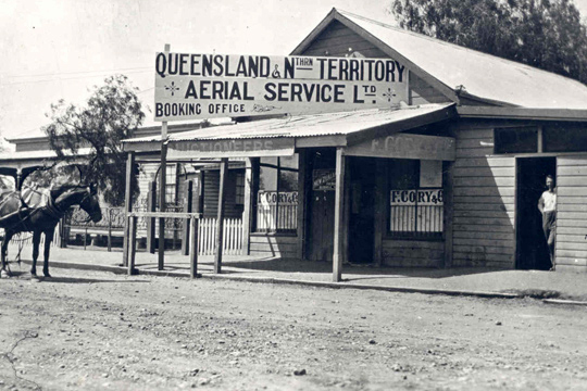 Timber shopfront with large sign on roof reading Queensland & Northern Territory Aerial Service Ltd Booking office. The road is unsealed and a horse in harness is visible at left. A man is standing is a doorway looking at the camera. - click to view larger image