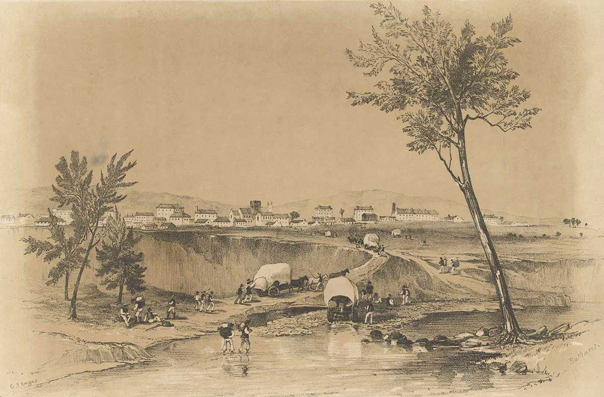 : lithograph showing a creek and a tree in the foreground with horse-drawn wagons. In the background is a small town made up of one- or two-storey white buildings.