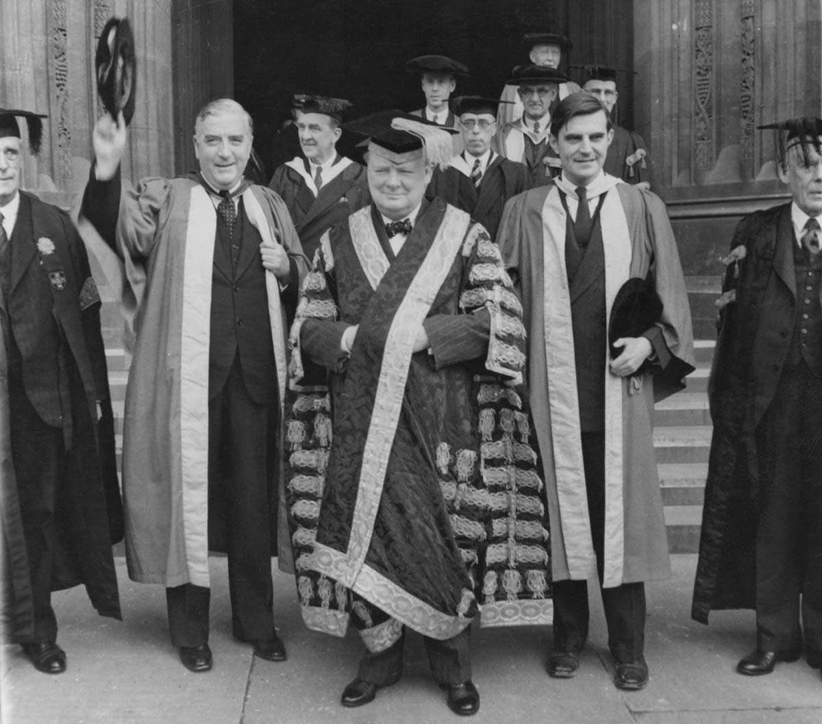 A black and white photo of a group of older men dressed in graduate garb.