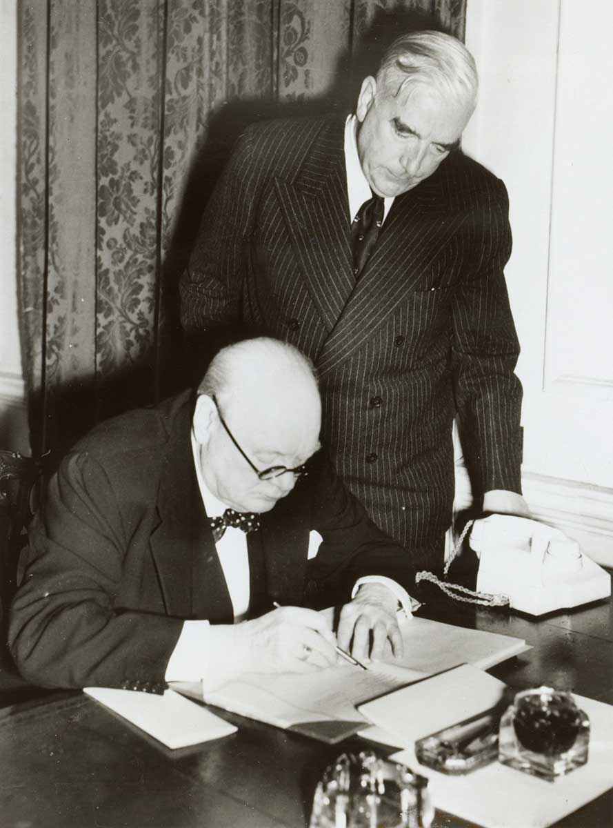 Churchill is at a desk examining some documents. Menzies is standing beside him, looking over his shoulder. - click to view larger image