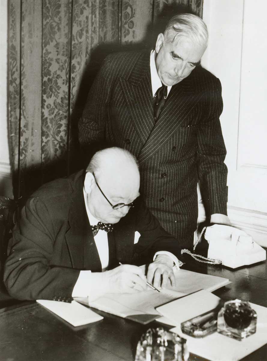 Churchill is at a desk examining some documents. Menzies is standing beside him, looking over his shoulder