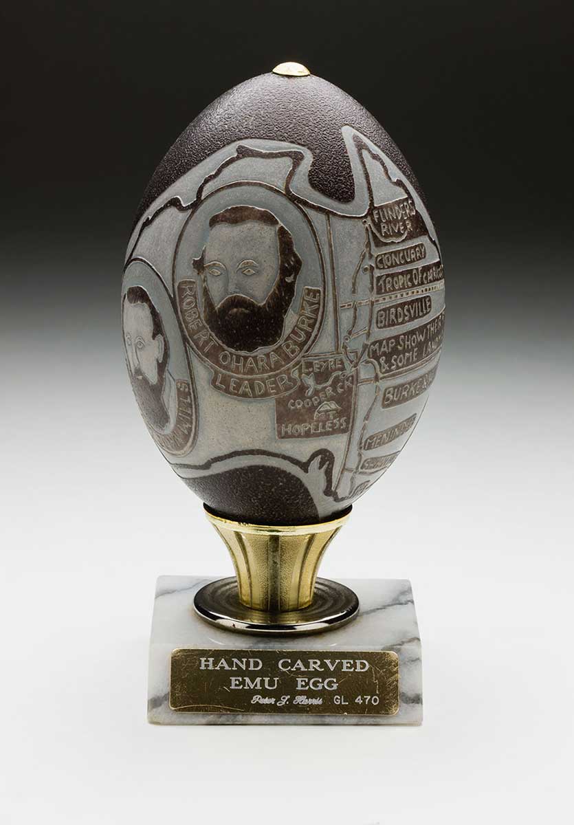 The egg, carved by craftsman Peter Harris, depicts a map of the journey of Burke and Wills. The egg rests on a marble base with a gold-plated plaque. The engraved text reads 'HAND CARVED / EMU EGG / Peter J. Harris GL 470'.