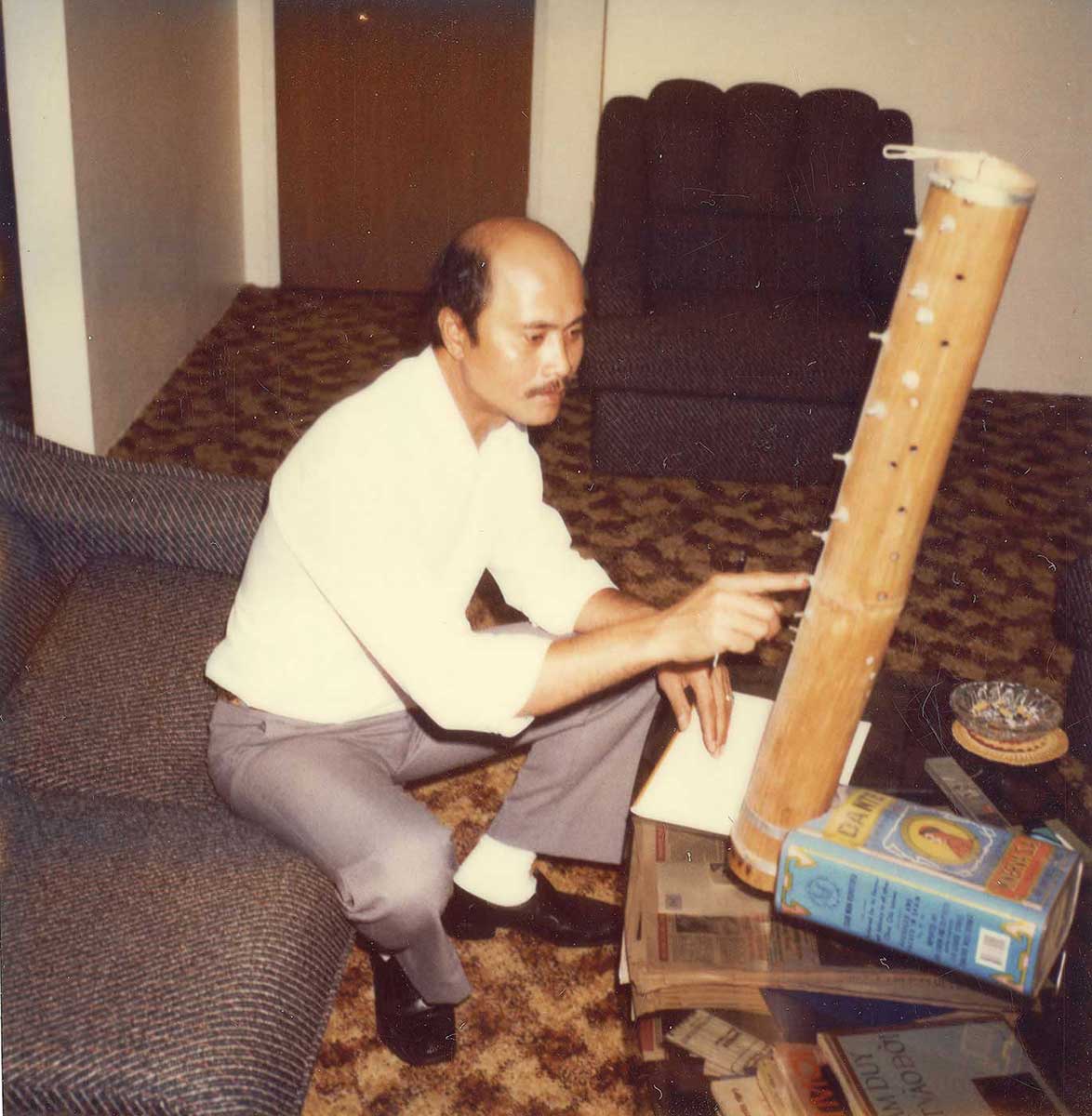 A man sits on a lounge. His right arm points to a bamboo musical instrument, which rests on a small table. The instrument has keys and strings down the side and an olive oil tin at the base. - click to view larger image
