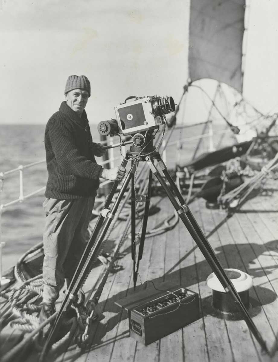Black and white photo of a man standing on a ship deck with a camera on a tripod. - click to view larger image
