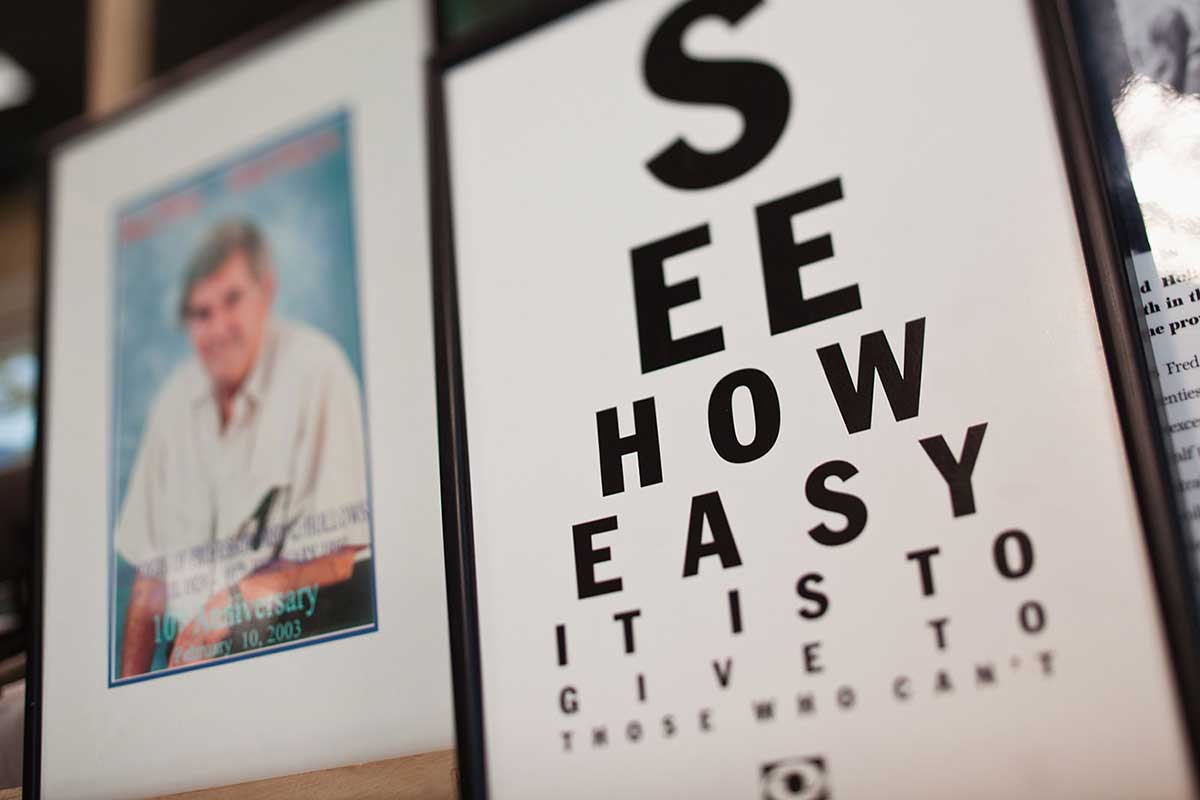 A poster at left in the style of an eye examination chart reads 'SEE HOW EASY IT IS TO GIVE TO THOSE WHO CAN'T'. A blurred portrait of Fred Hollows is on the right. - click to view larger image