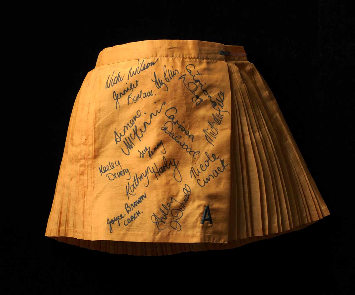 A golden yellow polyester netball skirt. The skirt is pleated, in alternating wide and narrow pleats. The front panel of the skirt is unpleated and signed in black ink by the members of the Australian women's netball team.