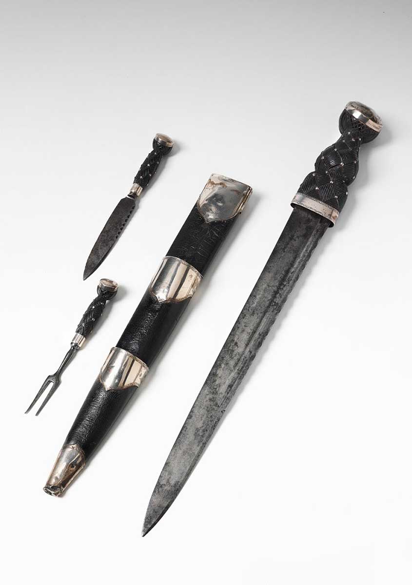 Dirk consisting of a large blade with a single edge. The back is notched in a decorative manner. The carved wooden hilt is studded with silver nails, and it has a plain silver pommel. It includes a small metal by-knife and fork with a carved wooden hilt and silver mounts. The leather scabbard has silver mounts. - click to view larger image