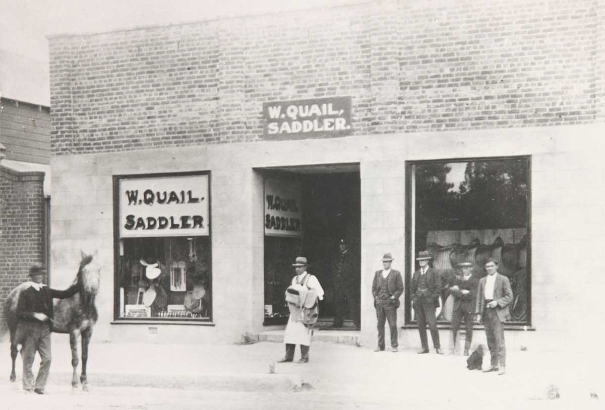 William Quail carrying a saddle outside his shop in Cooma to fit on a horse being held with four men standing outside the shop.