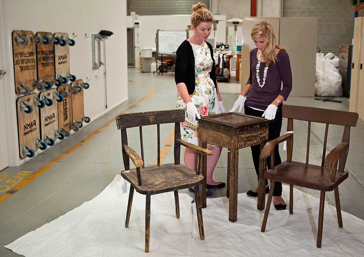 Two women stand behind a small wooden table and two chairs in a warehouse.