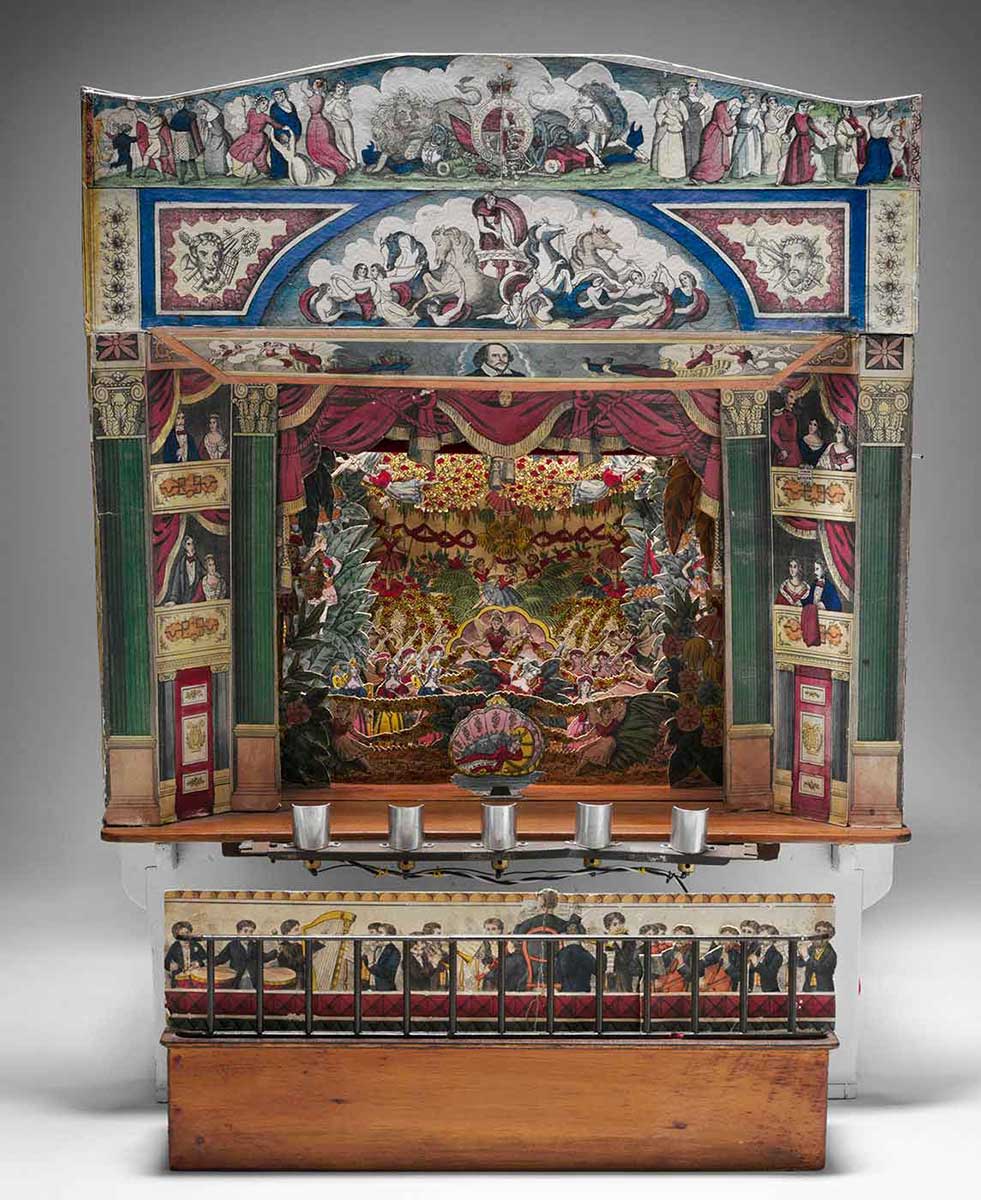 Front view of a brightly coloured toy stage, with cardboard sets and characters, and row of silver candle holders below. - click to view larger image