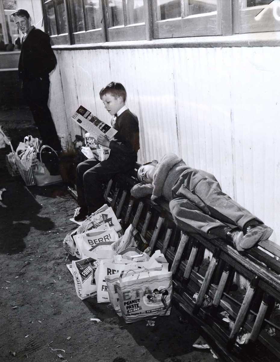 Black and white photograph showing two young boys, one lying with his eyes closed, on a ladder which rests longways, alongside a wall. Several show bags are lined up beneath him. Beside him, another boy sits reading. - click to view larger image