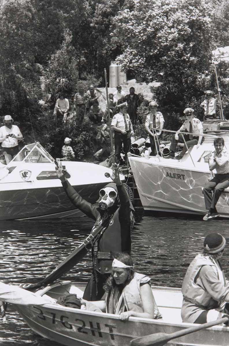 A man wearing a gas mask standing in a boat on a river. Police are observing the scene. - click to view larger image