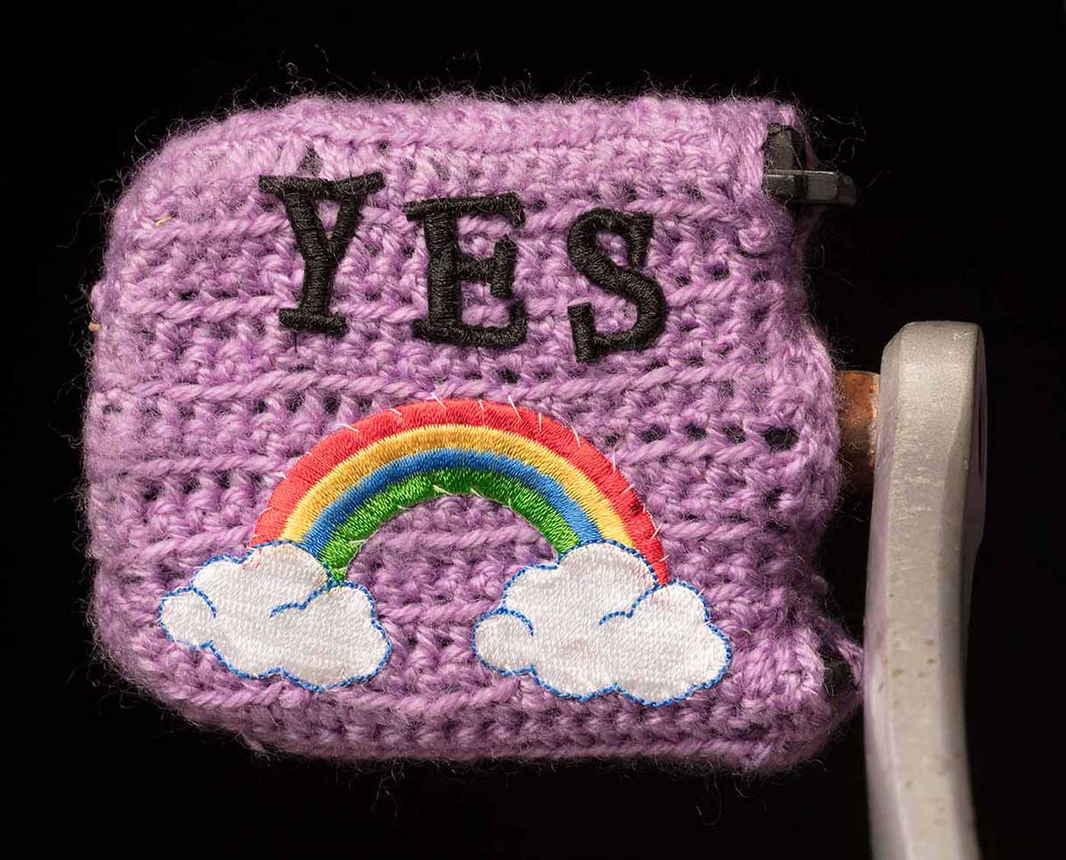 Studio photo of richly decorated pushbike pedal with the word 'YES' embroidered above an embroidered rainbow motif.