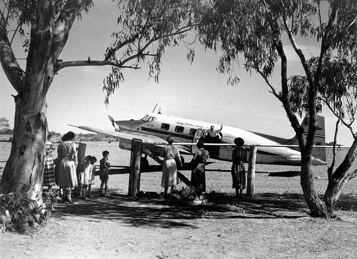 A black and white photo of a plane inside a paddock. There are several people, including children, on the other side of the fence facing the plane.