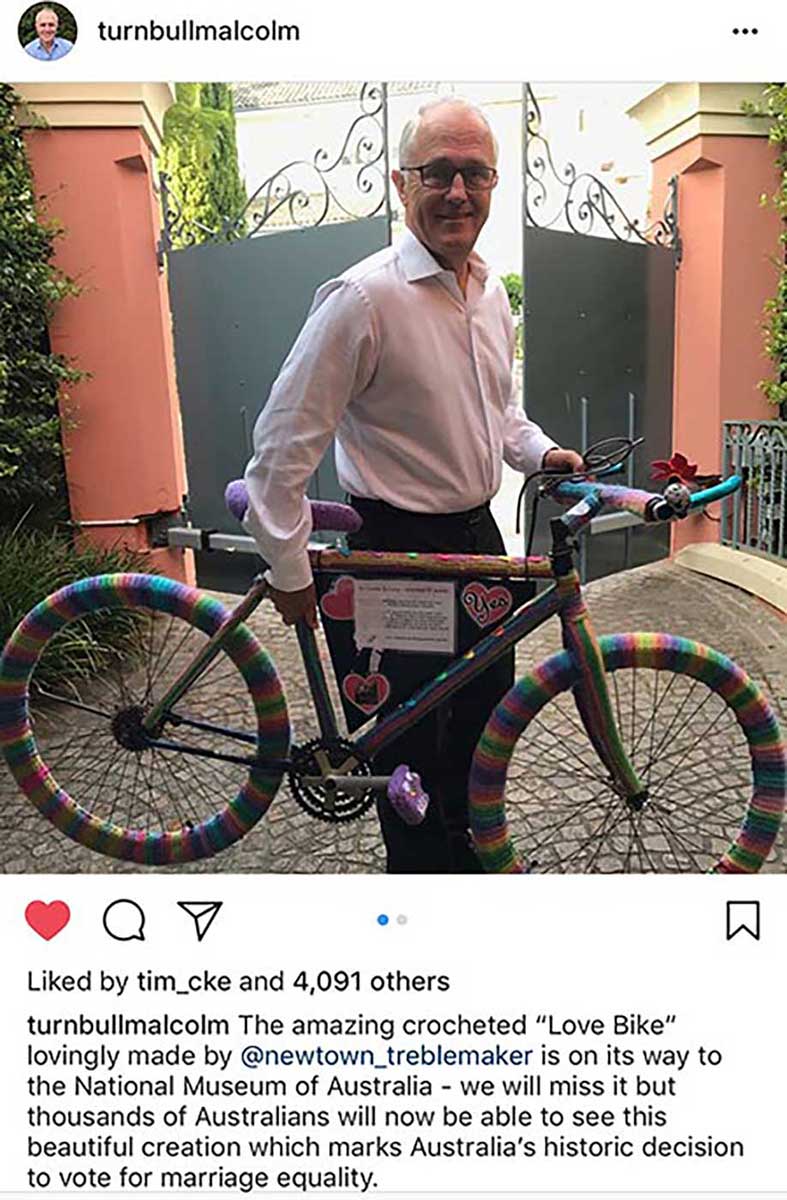 Man holding bike covered in rainbow-coloured wool. - click to view larger image