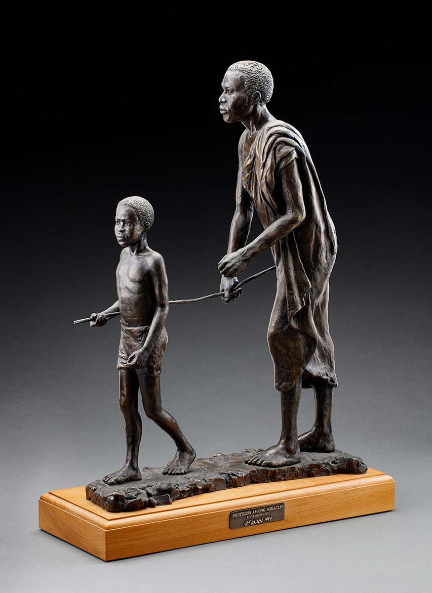 Statue of a boy in a loin cloth leading a man wearing robes. The boy holds a stick in his right hand. The other end of the stick is held by the man. - click to view larger image