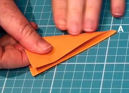 A piece of orange paper which has been partially folder over. Fingers from one hand hold down one side of the paper while the other hand holds a pencil which points to a corner with the capital letter A in white font.