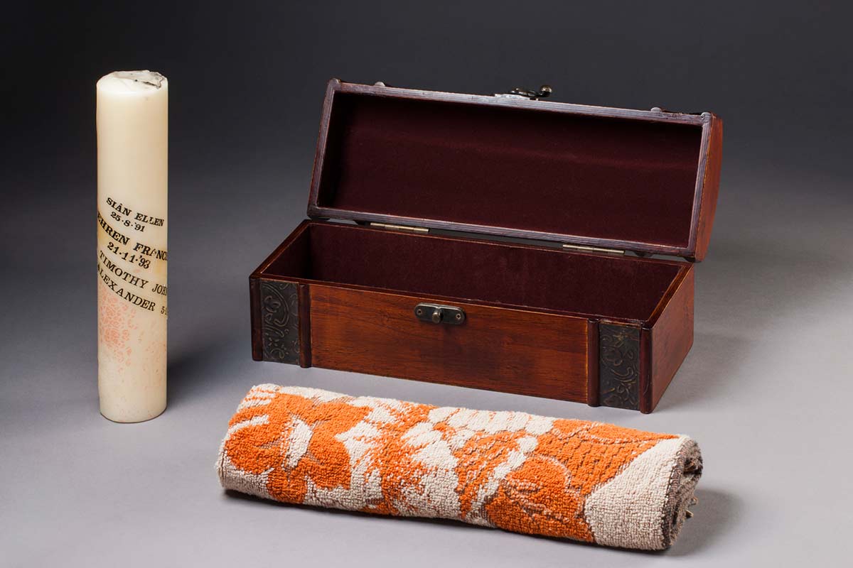 A lidded box containing a painted and inscribed candle and a small towel.