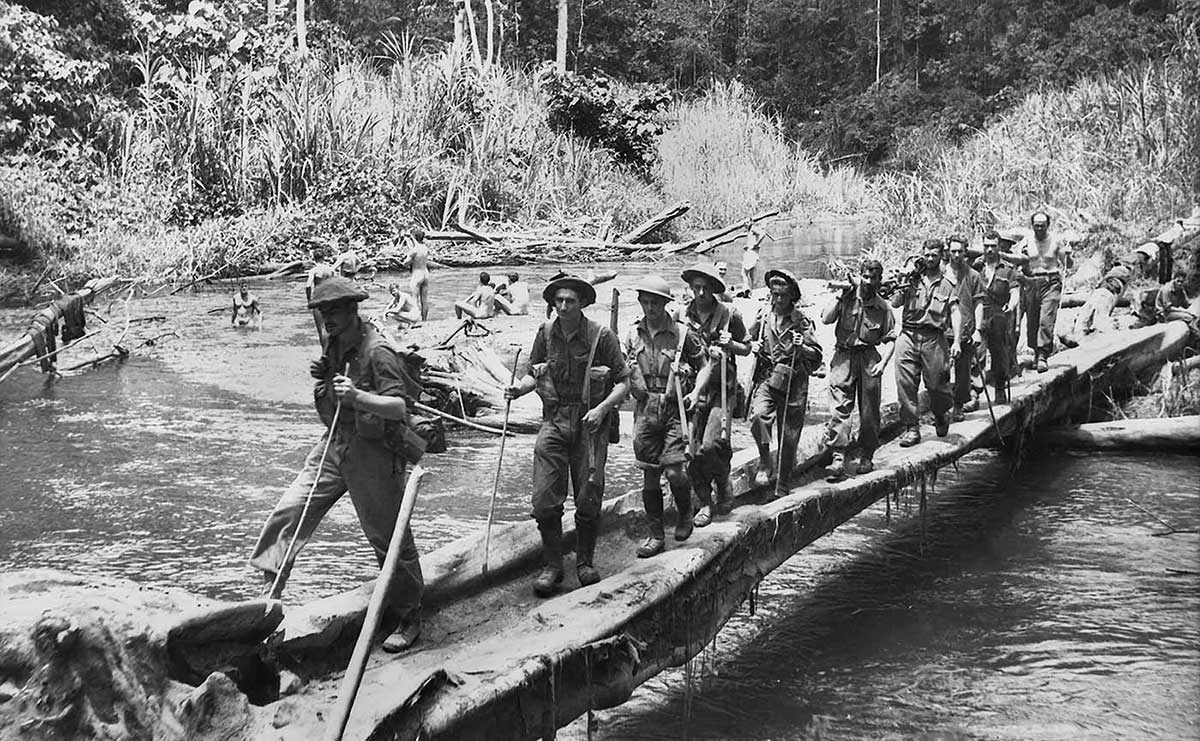Black and white photo of about a dozen soldiers walking across a rickety bridge over a river with tree-clad hills in background.