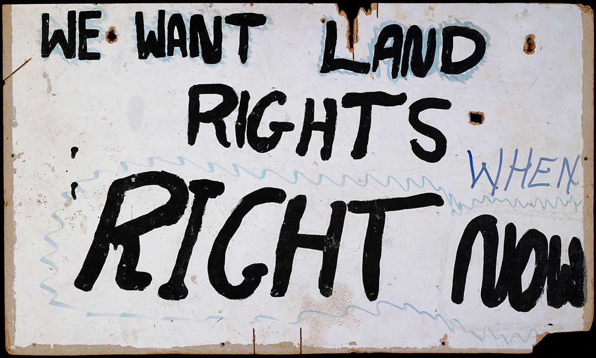 A placard. The text 'WE WANT LAND RIGHTS RIGHT NOW' has been painted in black paint on a rectangular masonite board, with a painted white background. 'WHEN' has been added in purple felt pen. - click to view larger image