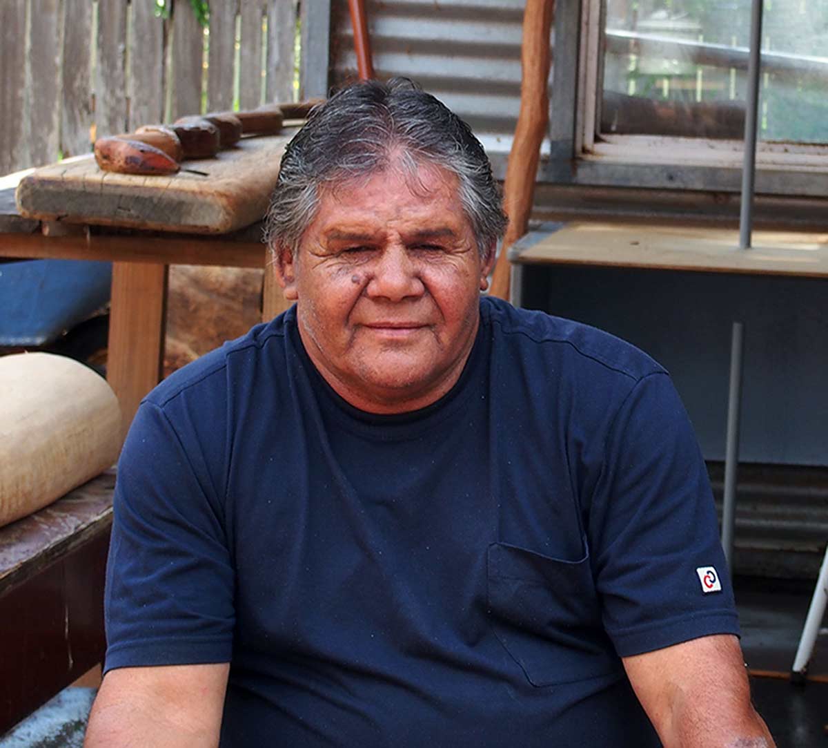 Portrait of an Indigenous Australian man seated in an outdoor workshop. - click to view larger image
