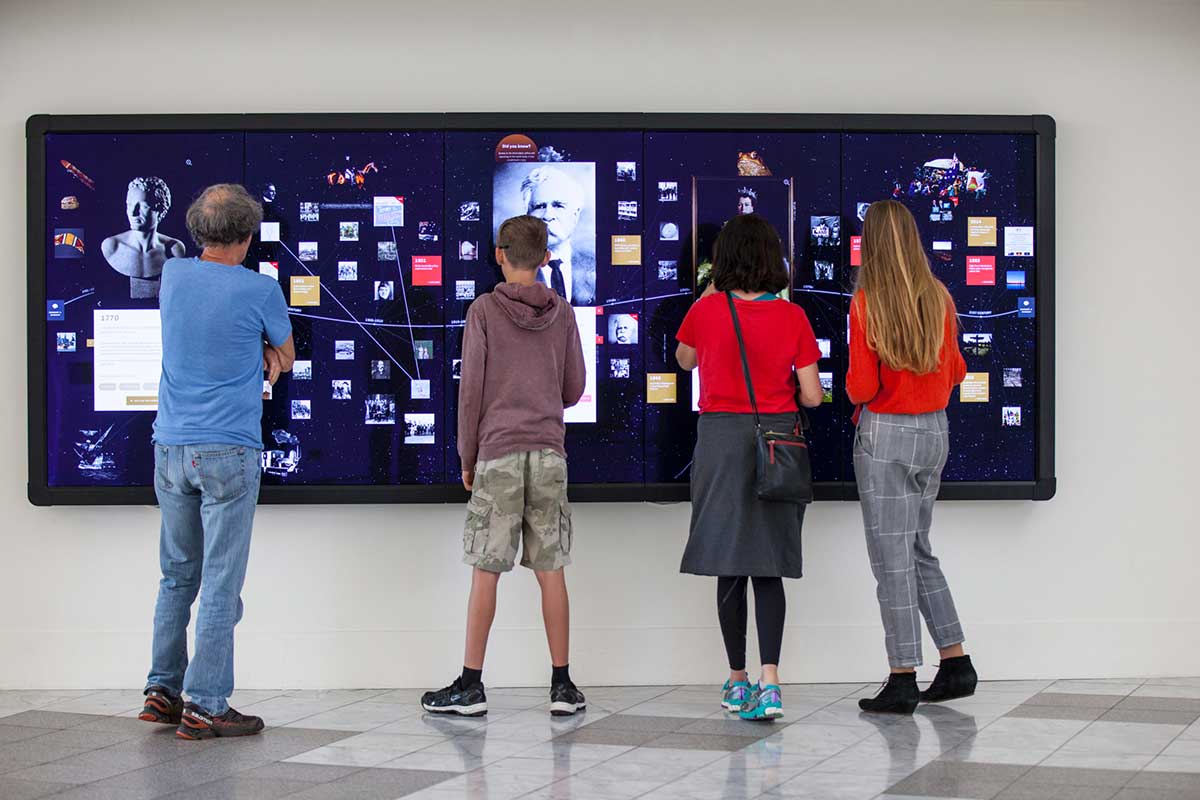 Four visitors interact with a large digital screen displaying a multitude of tiles with various content. 