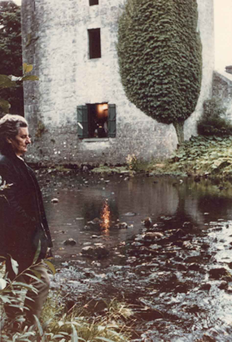 Faded photo of an elderly man standing by a large brook overlooked by a tall old stone building. - click to view larger image