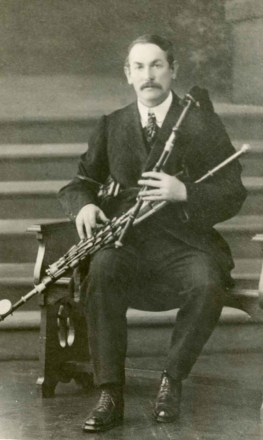 Black and white photo of a man sitting and holding an Irish musical pipe. - click to view larger image