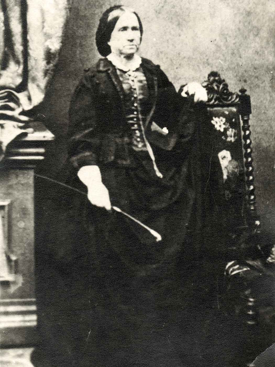 Black and white photo of a woman in black dress from the 19th Century and holding what appears to be a crop. - click to view larger image