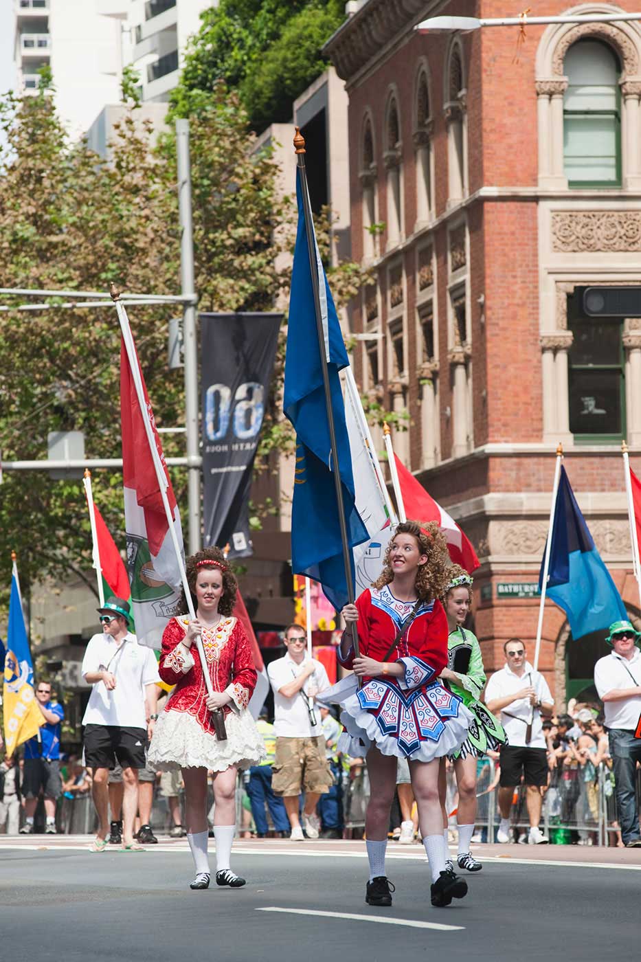 Procession of people including women dressed in traditional Irish dresses and carrying flags, while crowds look on. - click to view larger image
