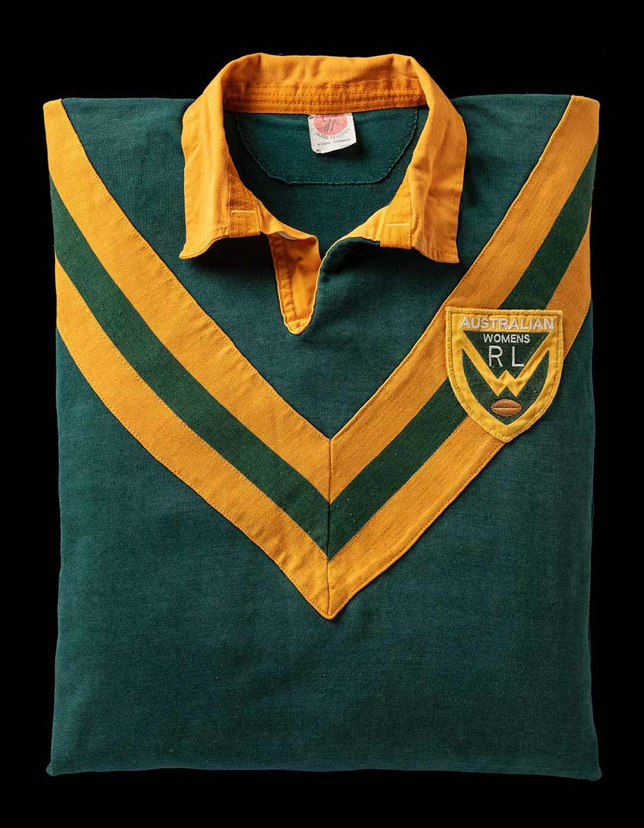 Front view of a neatly folded dark green rugby jersey with gold-coloured collar and two yellow v-shaped stripes which start from the shoulder and meet at the chest. A crest on the right breast has text 'Australian Womens RL'. - click to view larger image